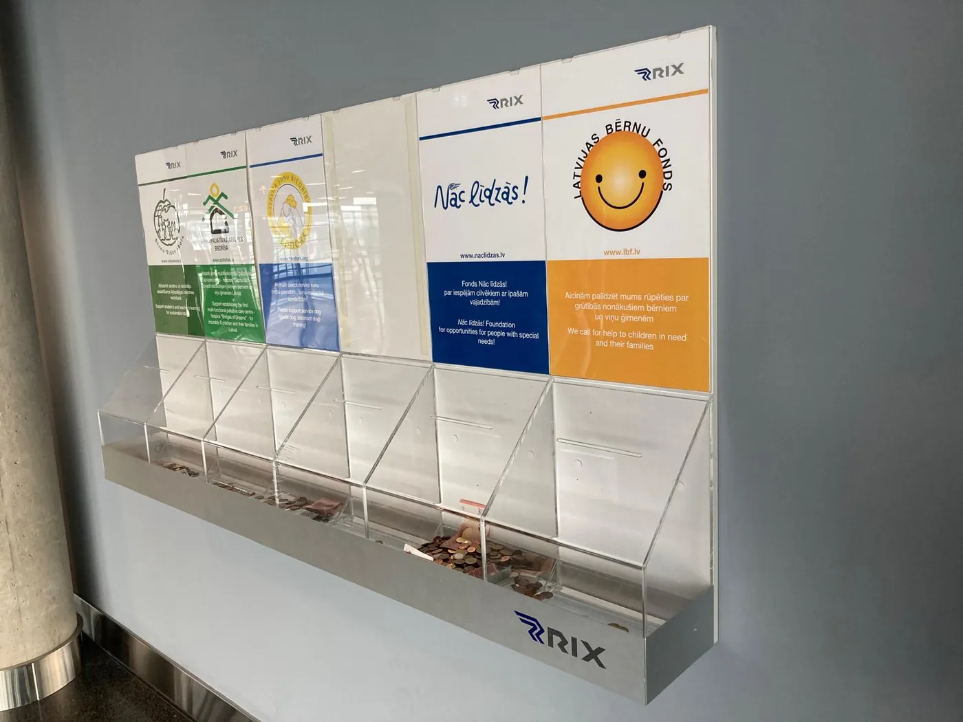 Row of clear collection boxes with printed charity panels, mounted on a wall at an airport