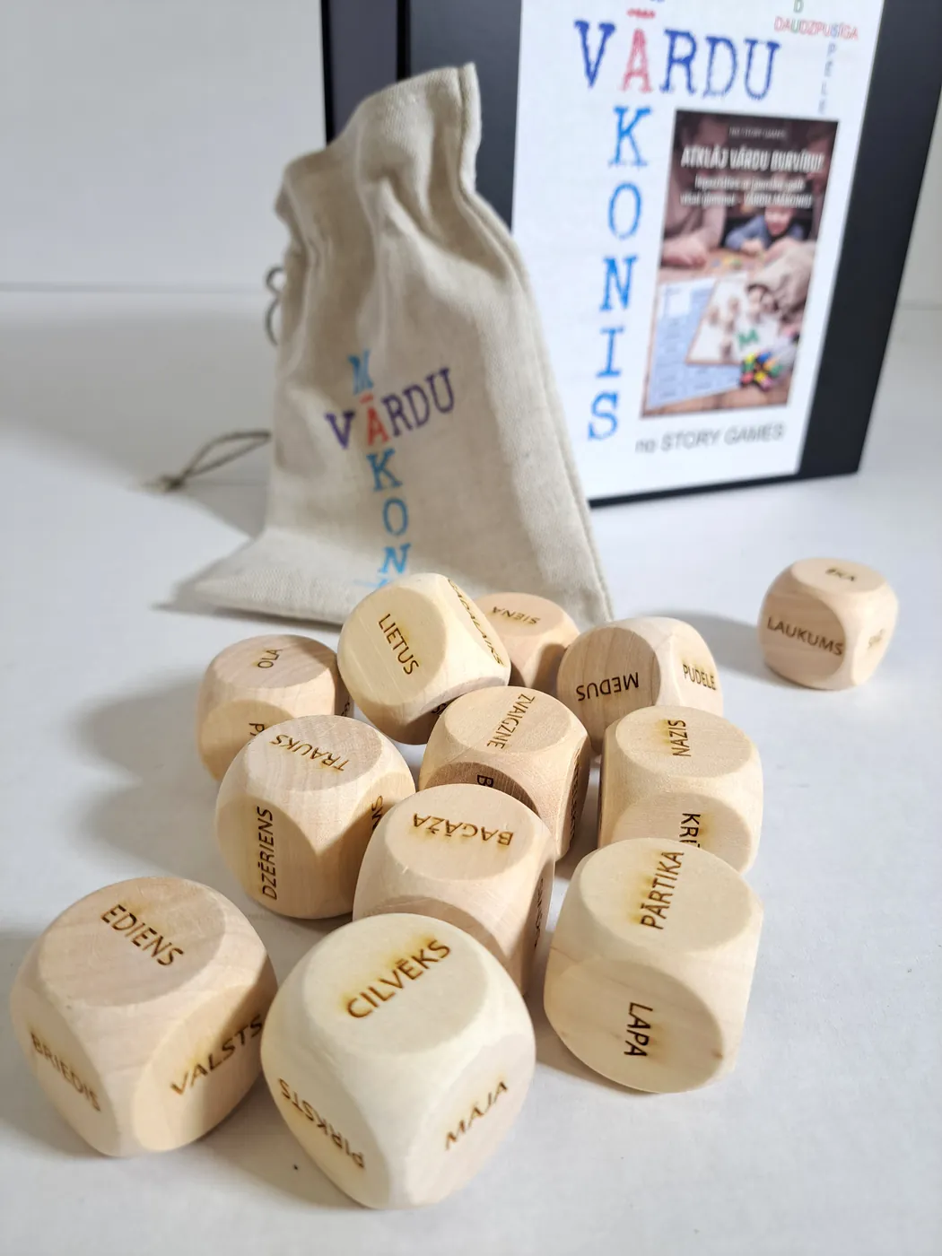 Wooden dice engraved with Latvian words, in front of a linen bag and the game's box