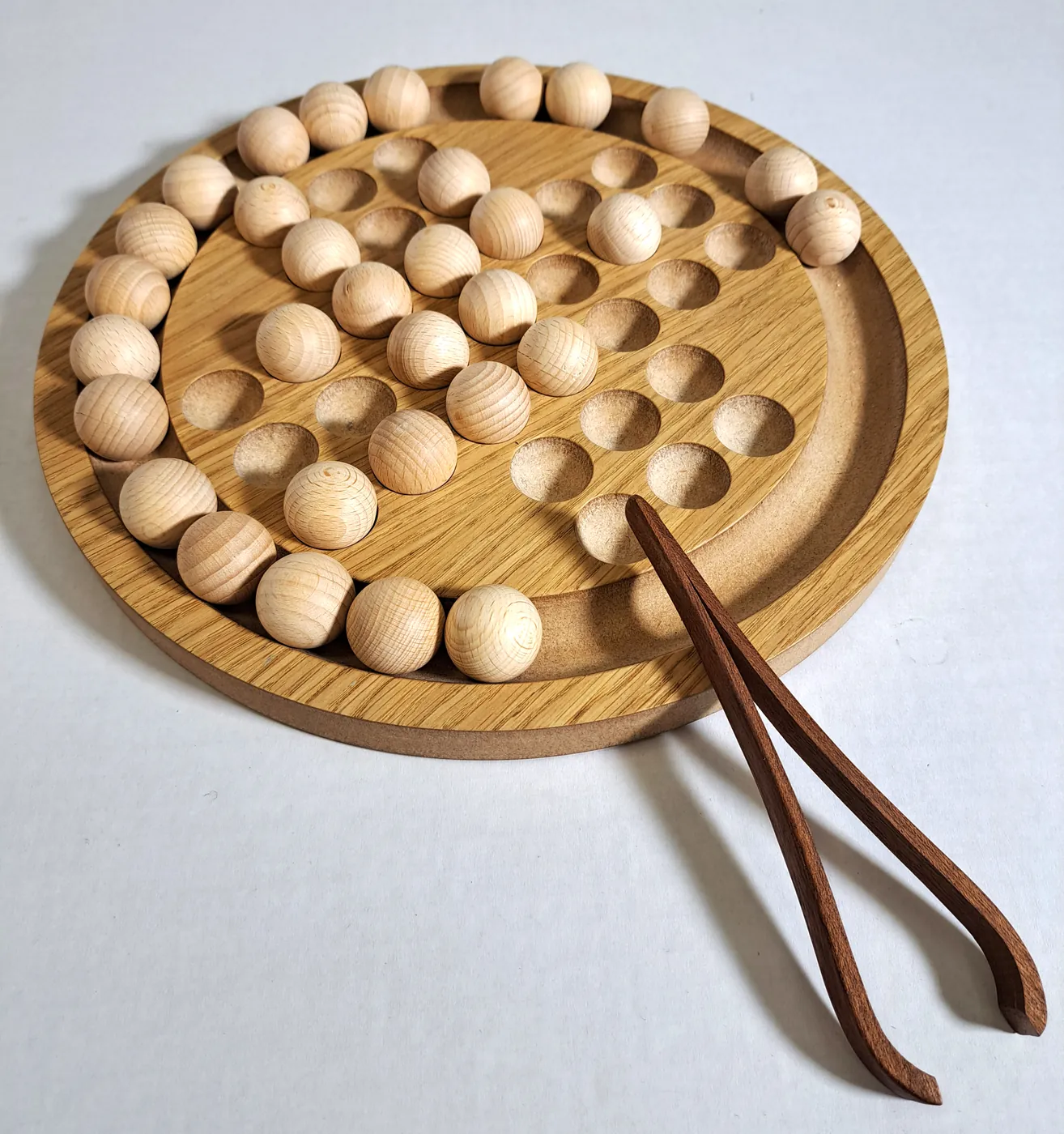 Round wooden solitaire board with a full set of wooden balls and a pair of wooden tongs