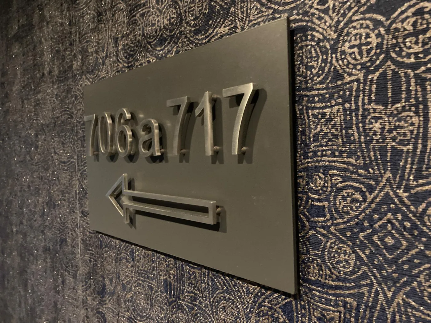 Dark plate with raised room numbers 706a to 717 and an outlined arrow, on a patterned corridor wall