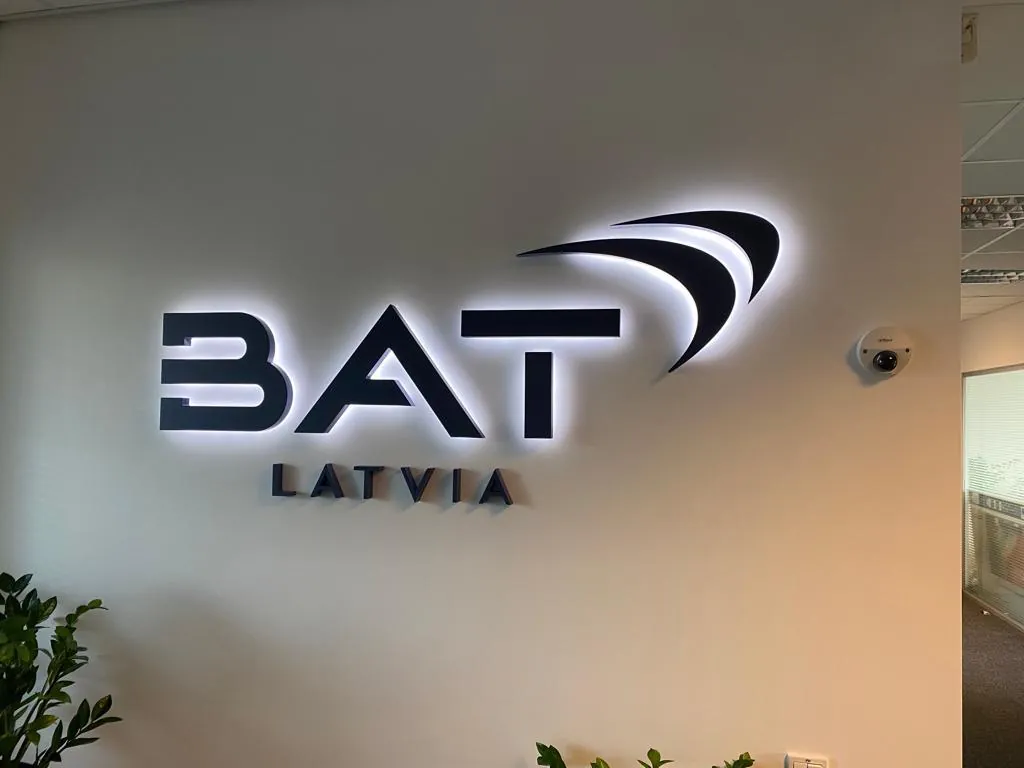 Dark logo letters on a light office wall, lit from behind so each letter casts a halo