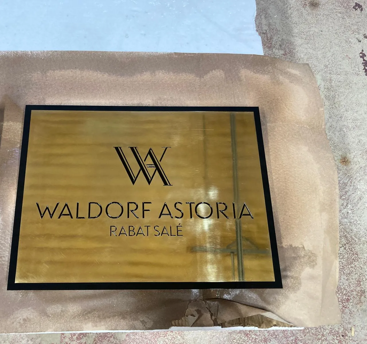 Gold-tone hotel plaque with a dark border and a logo, still in its protective wrapping