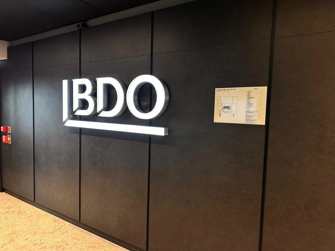 White illuminated logo letters with an underline bar, on a dark panelled office wall