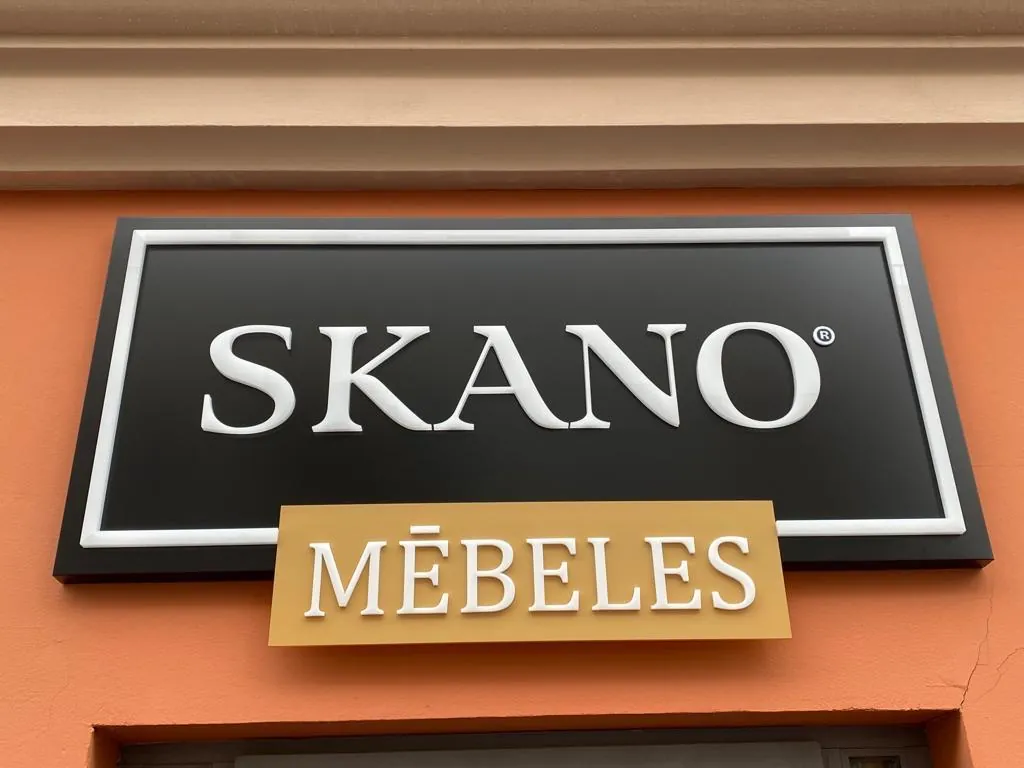 Shop sign on an orange facade: raised white letters on a black panel above a gold-tone plate