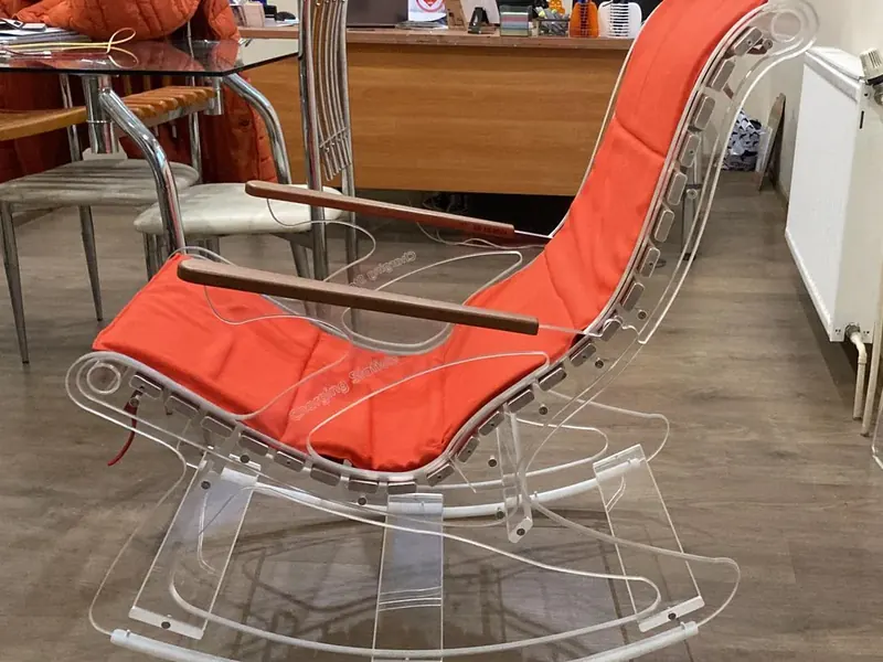 Rocking chair with a clear acrylic frame, an orange cushion and wooden armrests, seen from the side