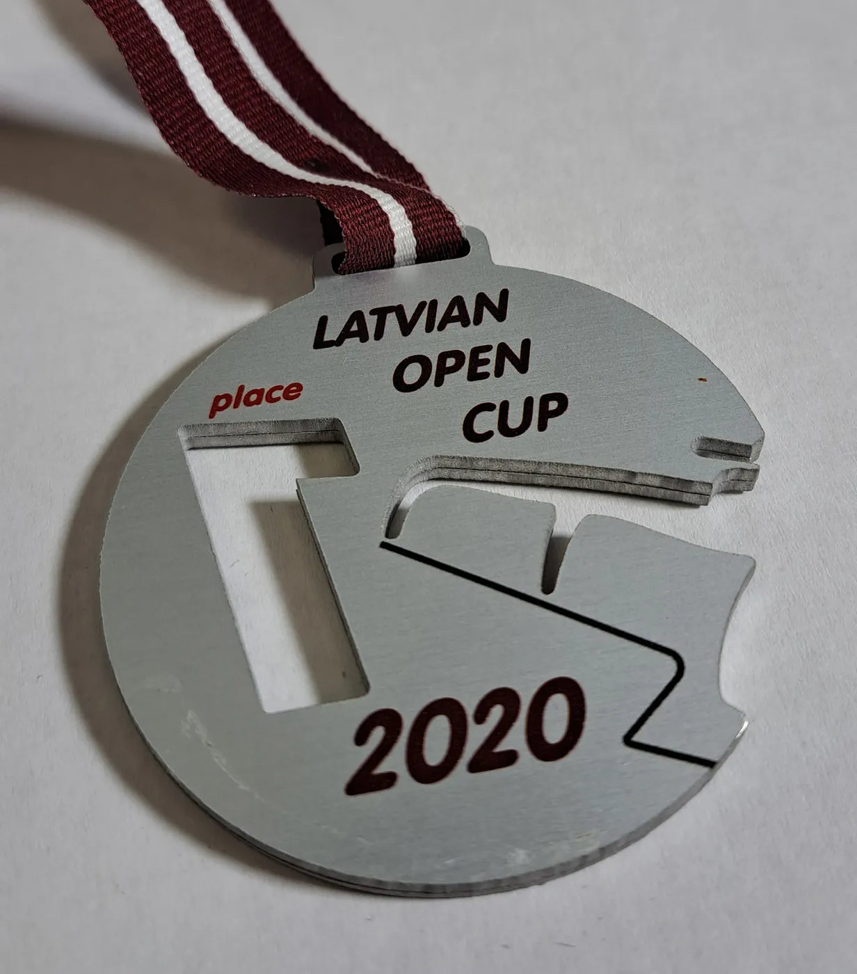 Brushed-metal medal for an open cup in 2020, with a cut-out track and a red and white ribbon